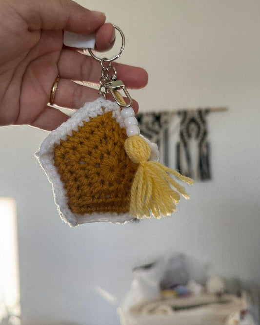 Hand holding a crochet house keychain with a yellow tassel against a neutral background